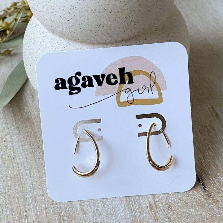 A pair of Agaveh Girl Gold Aspyn Hoops, featuring a minimalist, open curved design, displayed on a branded card with the text 'agaveh girl,' against a textured background. The earrings are dainty and modern, available in Ottawa, Ontario, Canada.
