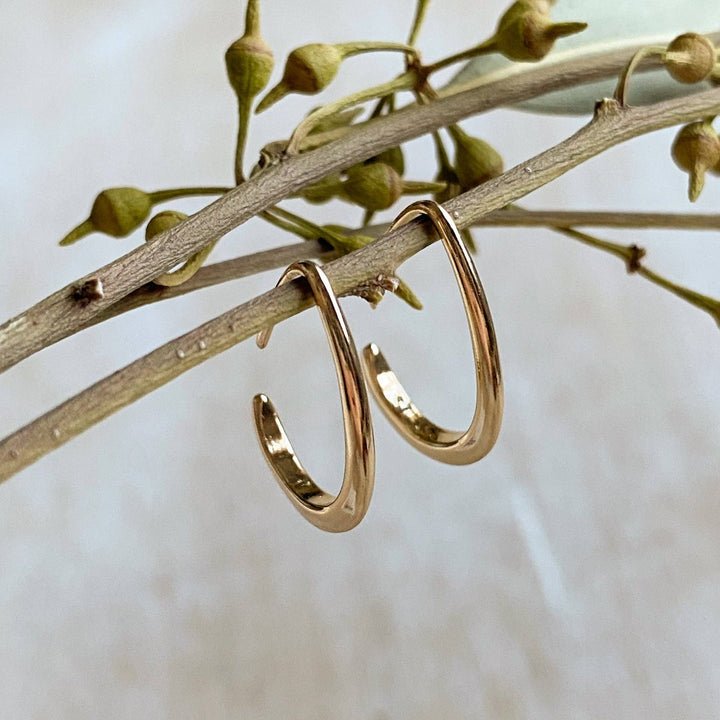 A pair of Agaveh Girl Gold Aspyn Hoops, featuring a minimalist, open curved design, displayed on a branded card with the text 'agaveh girl,' against a textured background. The earrings are dainty and modern, available in Ottawa, Ontario, Canada.