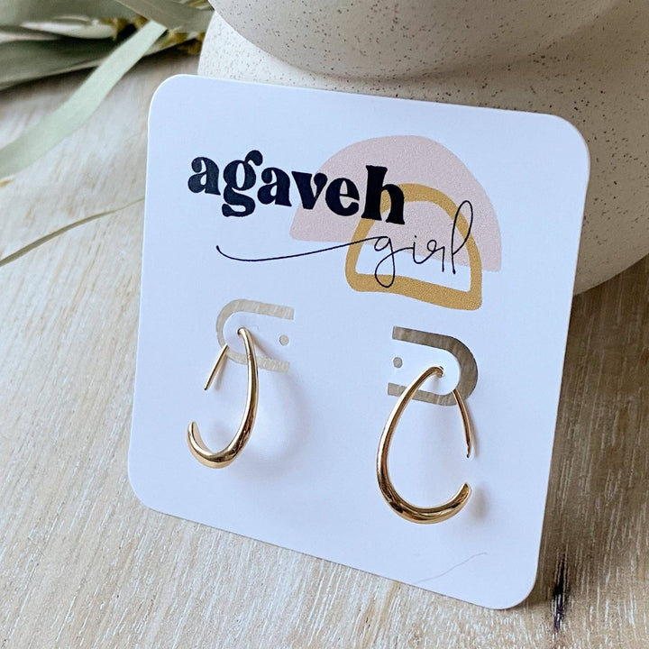 A pair of Agaveh Girl Gold Aspyn Hoops, featuring a minimalist, open curved design, displayed on a branded card with the text 'agaveh girl,' against a textured background. The earrings are dainty and modern, available in Ottawa, Ontario, Canada.