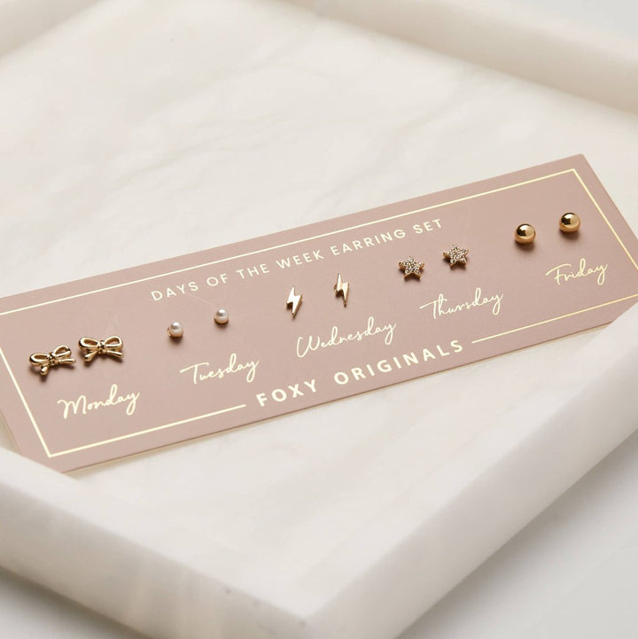 A set of gold stud earrings representing the days of the week. The earrings are arranged on a pink card with the words 