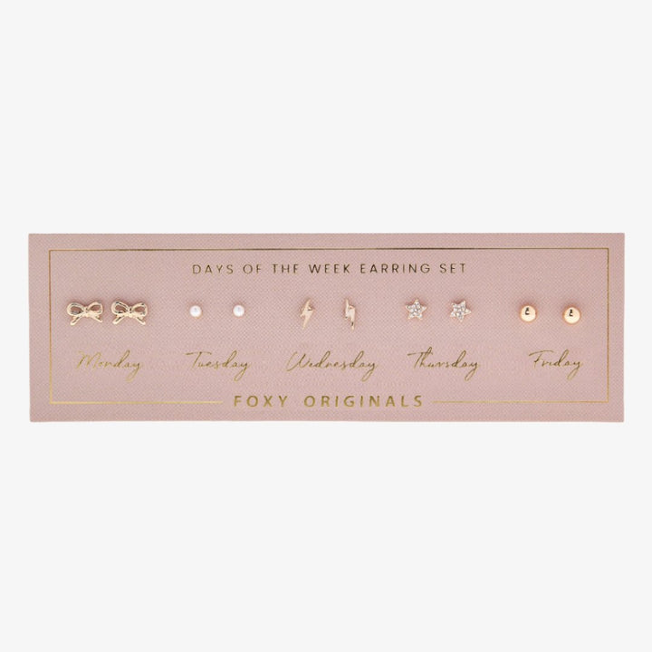 A set of gold stud earrings representing the days of the week. The earrings are arranged on a pink card with the words 