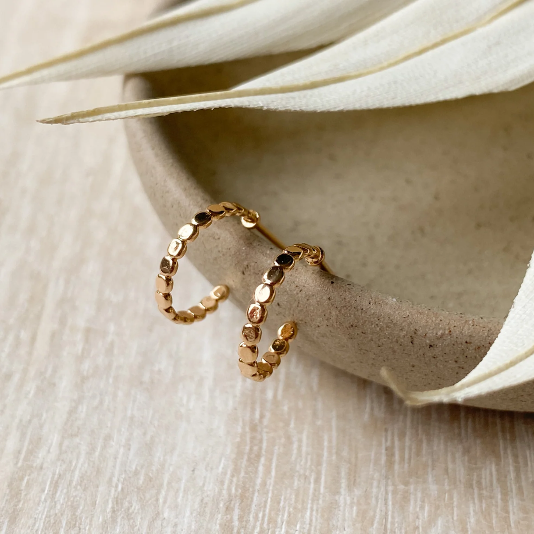 A pair of Agaveh Girl Delia Gold Plated Brass Hoops, featuring a textured, dotted design, displayed on a textured stone surface with dried floral accents. The earrings are dainty and gold, available in Ottawa, Ontario, Canada.