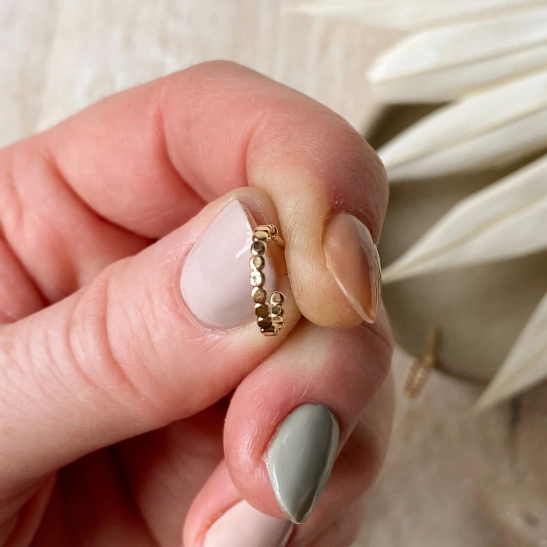 A pair of Agaveh Girl Delia Gold Plated Brass Hoops, featuring a textured, dotted design, displayed on a textured stone surface with dried floral accents. The earrings are dainty and gold, available in Ottawa, Ontario, Canada.