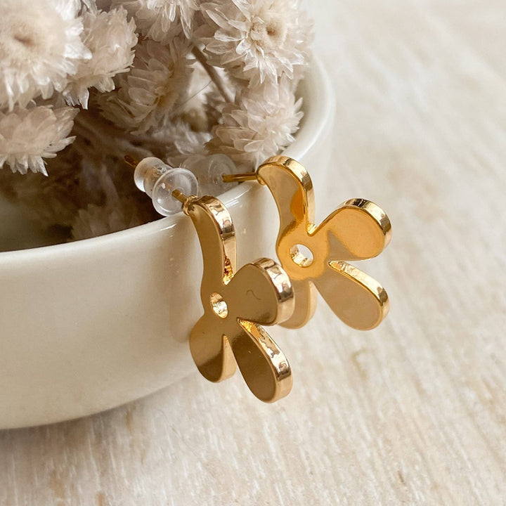 A pair of Agaveh Girl gold Forget Me Not Earrings, featuring a small, gold flower design with a beige center, displayed on a branded card with the text 'agaveh girl,' against a textured background with white floral accents. The earrings are dainty and floral, available in Ottawa, Ontario, Canada.
