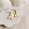 A pair of Agaveh Girl gold Forget Me Not Earrings, featuring a small, gold flower design with a beige center, displayed on a branded card with the text 'agaveh girl,' against a textured background with white floral accents. The earrings are dainty and floral, available in Ottawa, Ontario, Canada.