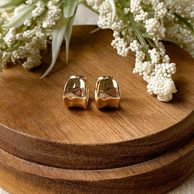 A pair of gold Grace hoops, featuring a chunky, domed design, displayed on a textured wooden surface with white floral accents. The earrings are statement huggie hoops, available in Ottawa, Ontario, Canada.