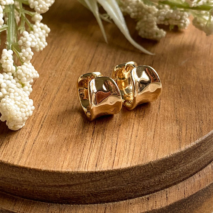 A pair of gold Grace hoops, featuring a chunky, domed design, displayed on a textured wooden surface with white floral accents. The earrings are statement huggie hoops, available in Ottawa, Ontario, Canada.