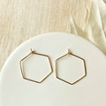 A pair of Agaveh Girl Gold Hexi Hoops, featuring a thin, gold wire hexagon design, displayed on a branded card with the text 'agaveh girl,' against a textured background with dried floral accents. The earrings are minimalist and geometric, available in Ottawa, Ontario, Canada.