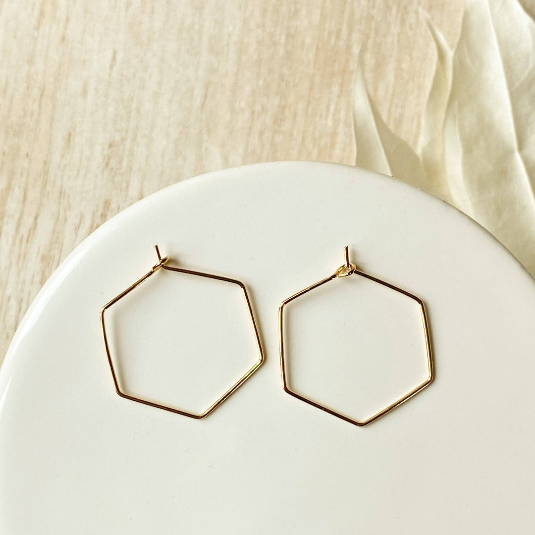 A pair of Agaveh Girl Gold Hexi Hoops, featuring a thin, gold wire hexagon design, displayed on a branded card with the text 'agaveh girl,' against a textured background with dried floral accents. The earrings are minimalist and geometric, available in Ottawa, Ontario, Canada.