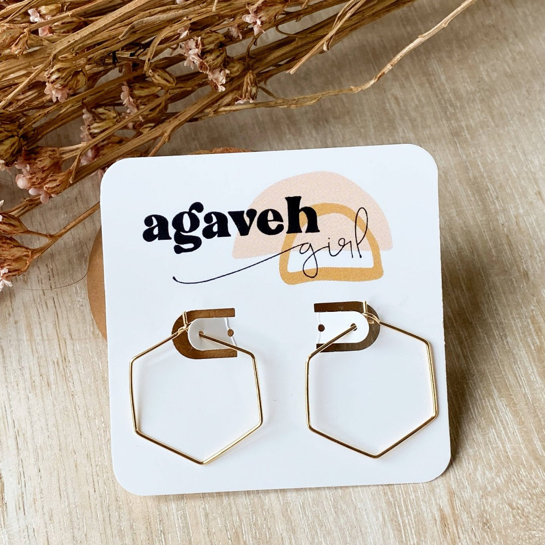 A pair of Agaveh Girl Gold Hexi Hoops, featuring a thin, gold wire hexagon design, displayed on a branded card with the text 'agaveh girl,' against a textured background with dried floral accents. The earrings are minimalist and geometric, available in Ottawa, Ontario, Canada.