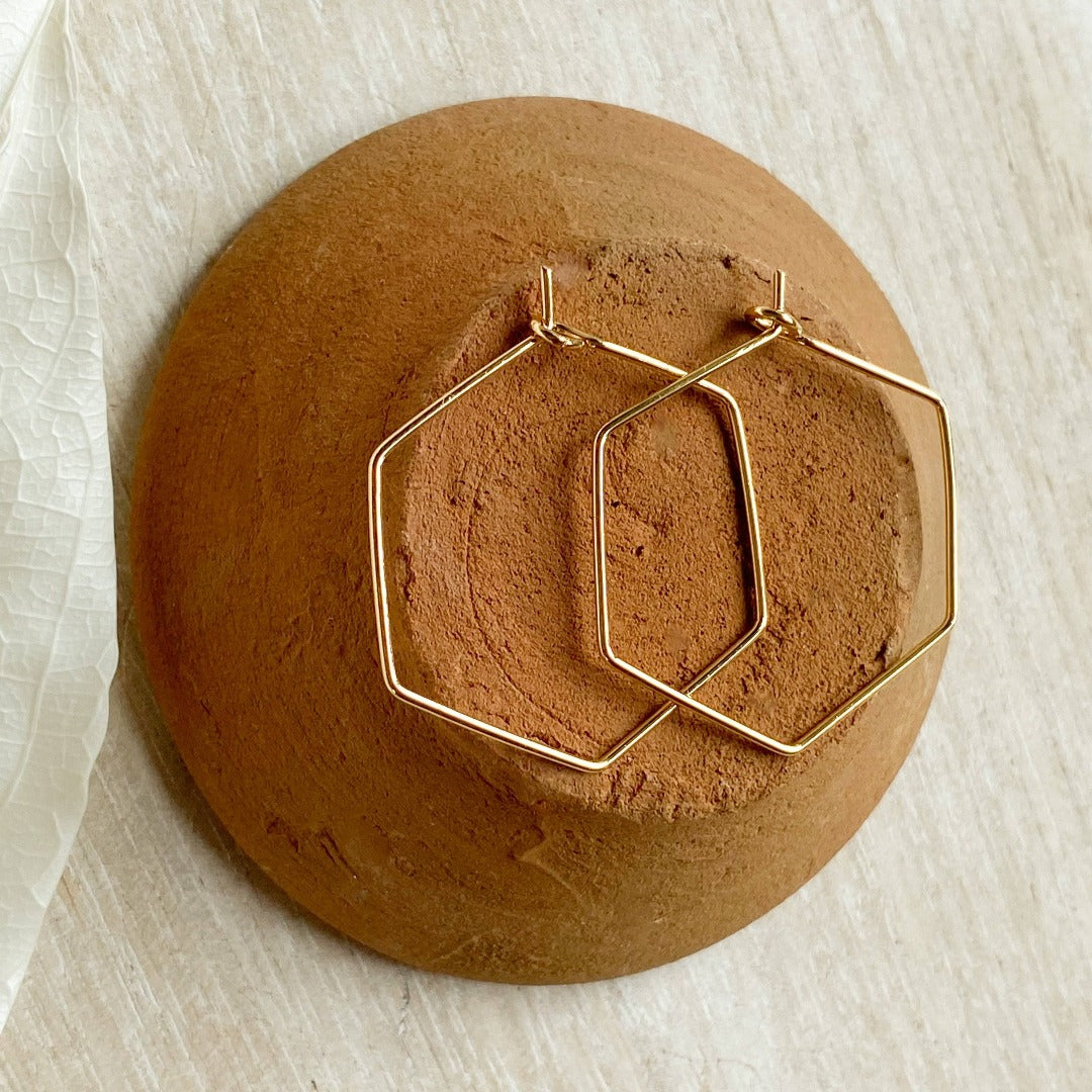 A pair of Agaveh Girl Gold Hexi Hoops, featuring a thin, gold wire hexagon design, displayed on a branded card with the text 'agaveh girl,' against a textured background with dried floral accents. The earrings are minimalist and geometric, available in Ottawa, Ontario, Canada.