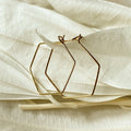 A pair of Agaveh Girl Gold Hexi Hoops, featuring a thin, gold wire hexagon design, displayed on a branded card with the text 'agaveh girl,' against a textured background with dried floral accents. The earrings are minimalist and geometric, available in Ottawa, Ontario, Canada.