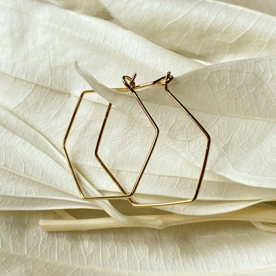 A pair of Agaveh Girl Gold Hexi Hoops, featuring a thin, gold wire hexagon design, displayed on a branded card with the text 'agaveh girl,' against a textured background with dried floral accents. The earrings are minimalist and geometric, available in Ottawa, Ontario, Canada.