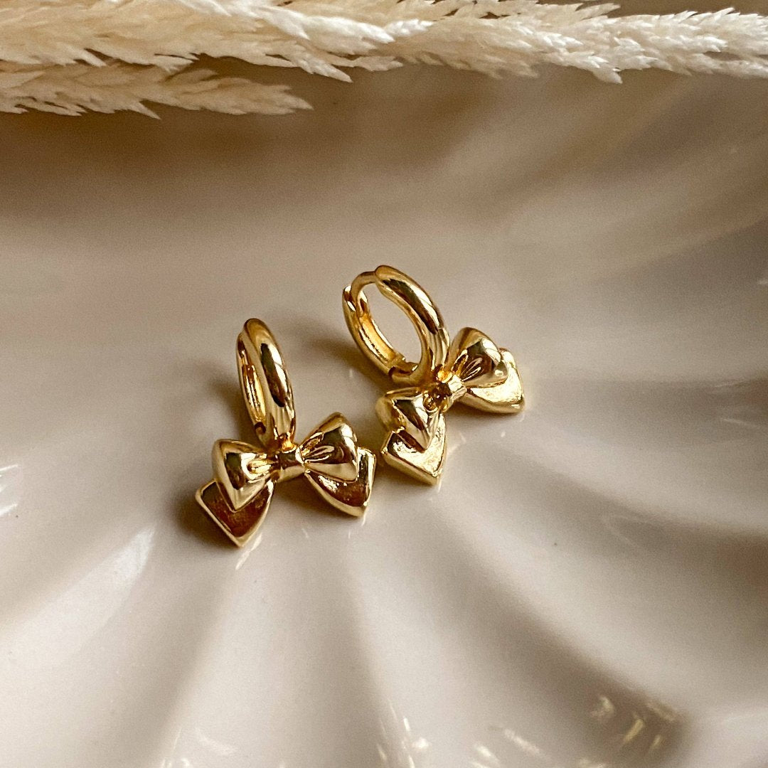 A pair of gold Jude hoops, featuring a dainty bow huggie design, displayed on a scalloped ceramic dish with dried floral accents. The earrings are delicate and feminine, available in Ottawa, Ontario, Canada.