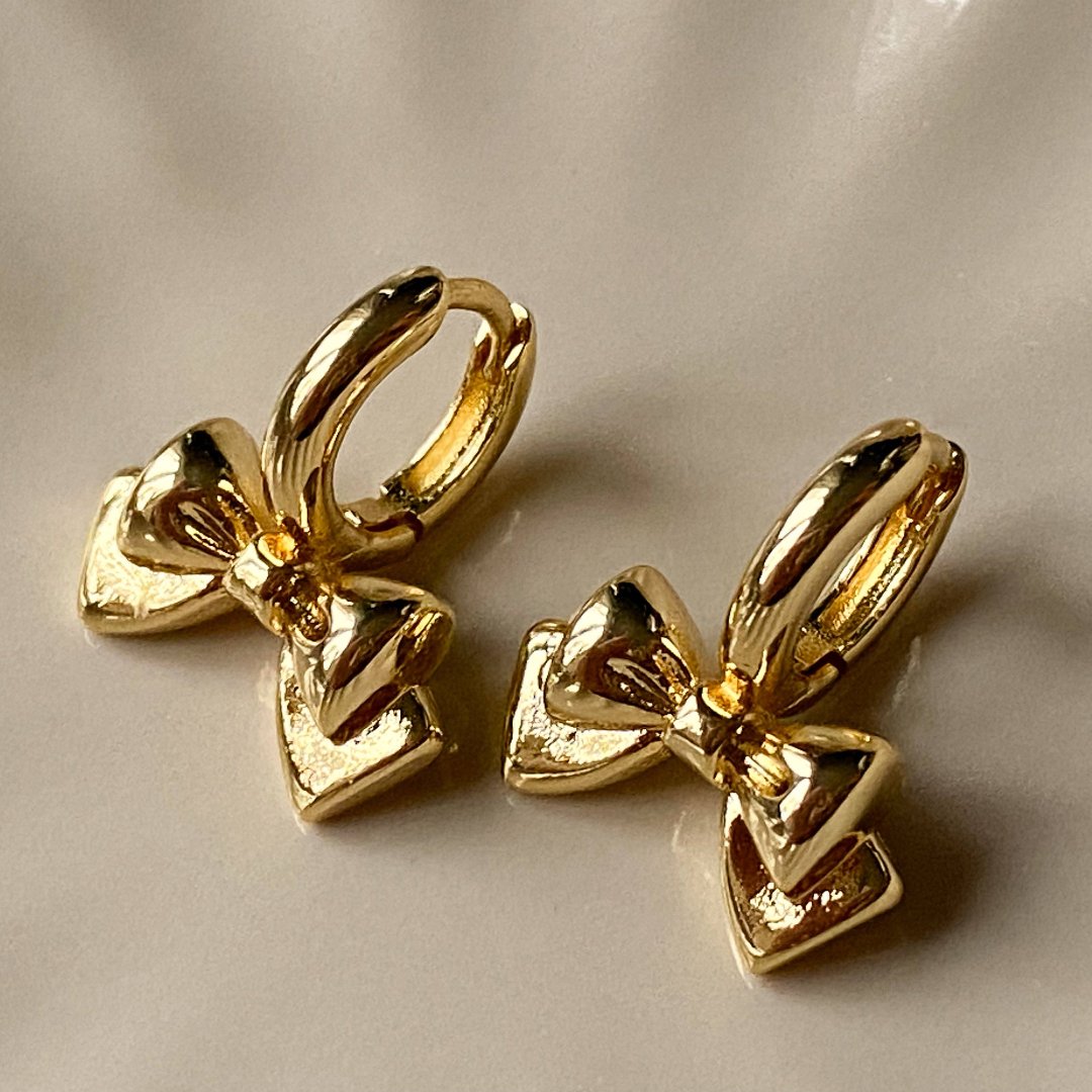 A pair of gold Jude hoops, featuring a dainty bow huggie design, displayed on a scalloped ceramic dish with dried floral accents. The earrings are delicate and feminine, available in Ottawa, Ontario, Canada.