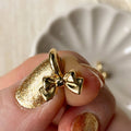 A pair of gold Jude hoops, featuring a dainty bow huggie design, displayed on a scalloped ceramic dish with dried floral accents. The earrings are delicate and feminine, available in Ottawa, Ontario, Canada.