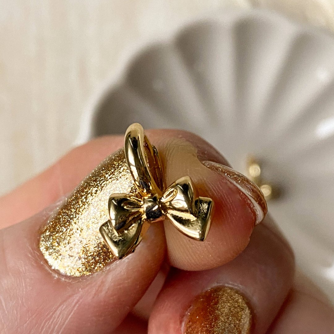 A pair of gold Jude hoops, featuring a dainty bow huggie design, displayed on a scalloped ceramic dish with dried floral accents. The earrings are delicate and feminine, available in Ottawa, Ontario, Canada.
