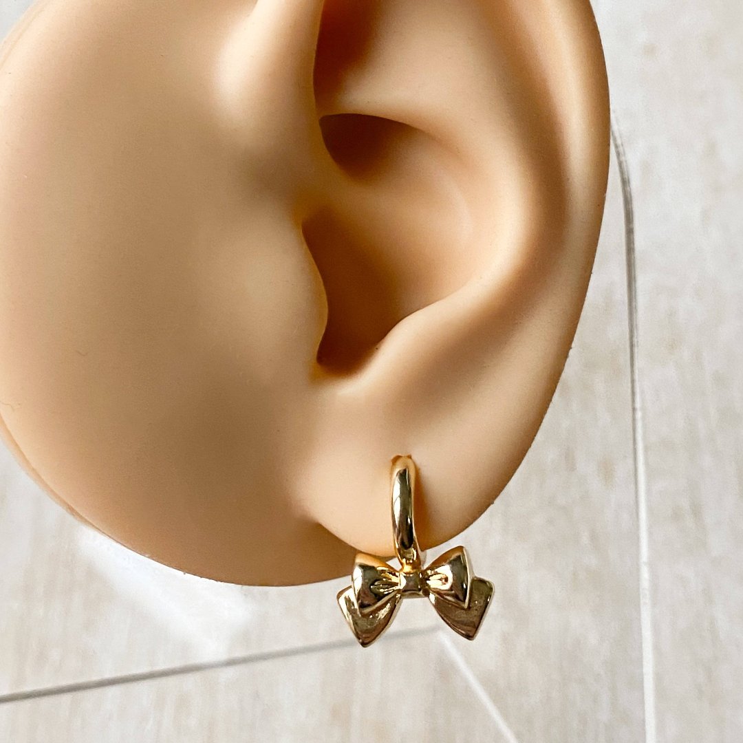 A pair of gold Jude hoops, featuring a dainty bow huggie design, displayed on a scalloped ceramic dish with dried floral accents. The earrings are delicate and feminine, available in Ottawa, Ontario, Canada.
