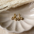 A pair of gold Jude hoops, featuring a dainty bow huggie design, displayed on a scalloped ceramic dish with dried floral accents. The earrings are delicate and feminine, available in Ottawa, Ontario, Canada.