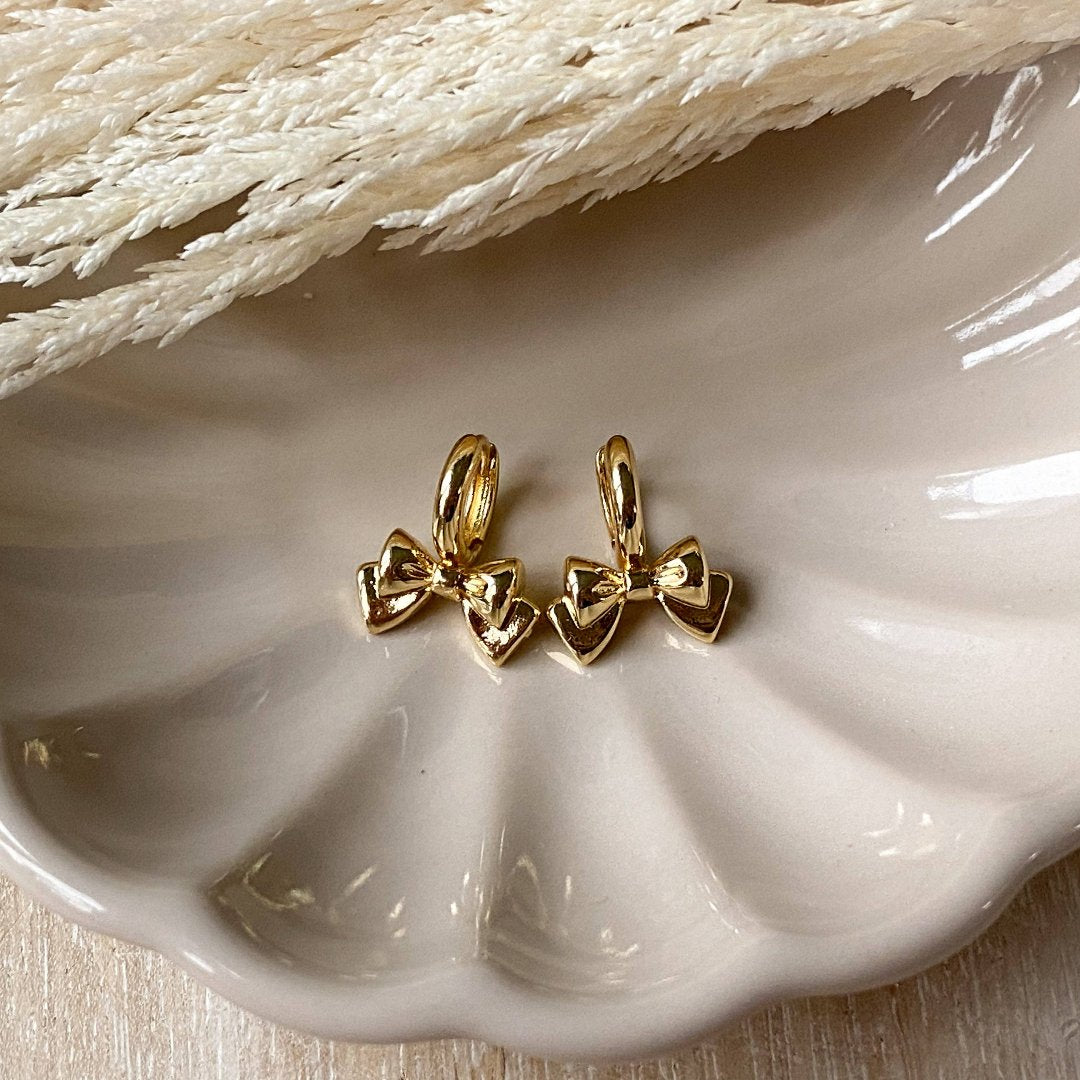 A pair of gold Jude hoops, featuring a dainty bow huggie design, displayed on a scalloped ceramic dish with dried floral accents. The earrings are delicate and feminine, available in Ottawa, Ontario, Canada.