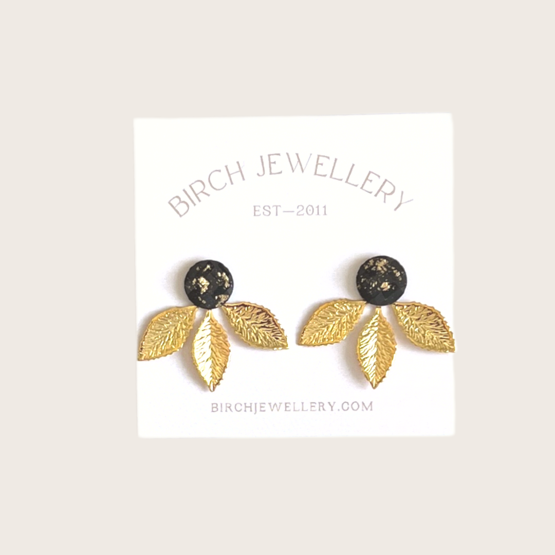 A pair of gold earrings with a leaf design. The earrings have a black, circular resin center with gold flecks. The leaves are attached to the center and can be detached.