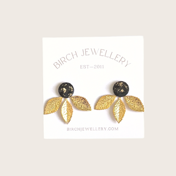 A pair of gold earrings with a leaf design. The earrings have a black, circular resin center with gold flecks. The leaves are attached to the center and can be detached.