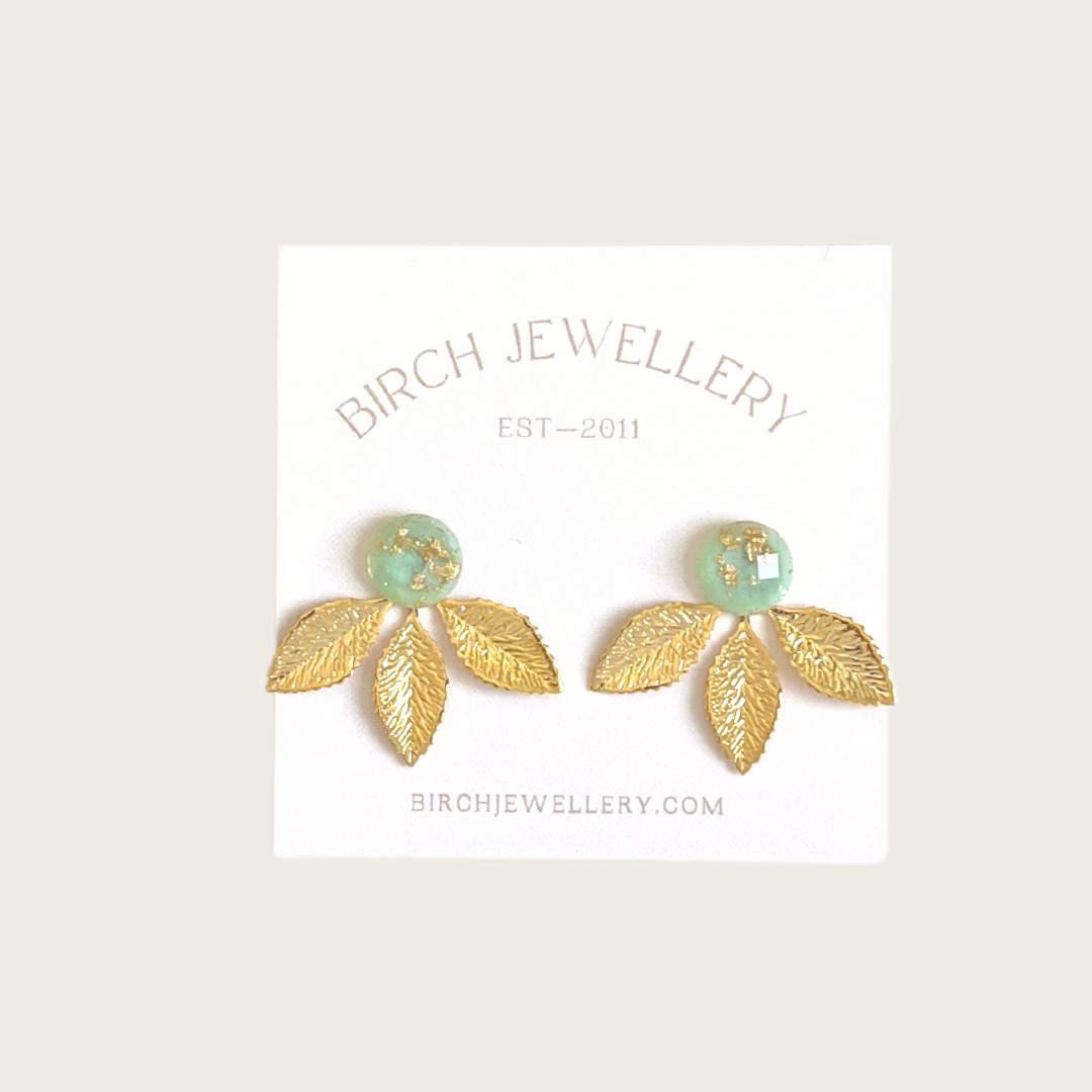 A pair of gold earrings with a leaf design. The earrings have a green, circular resin center with gold flecks. The leaves are attached to the center and can be detached.