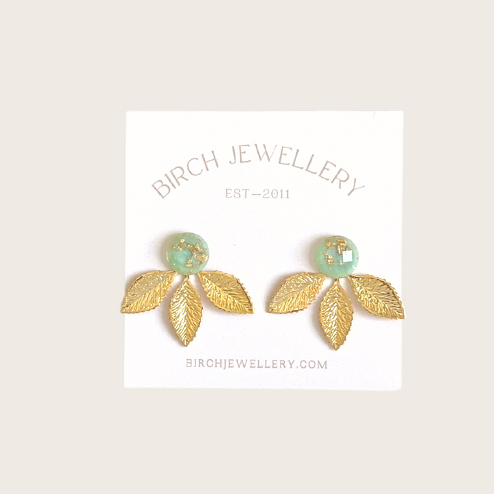 A pair of gold earrings with a leaf design. The earrings have a green, circular resin center with gold flecks. The leaves are attached to the center and can be detached.