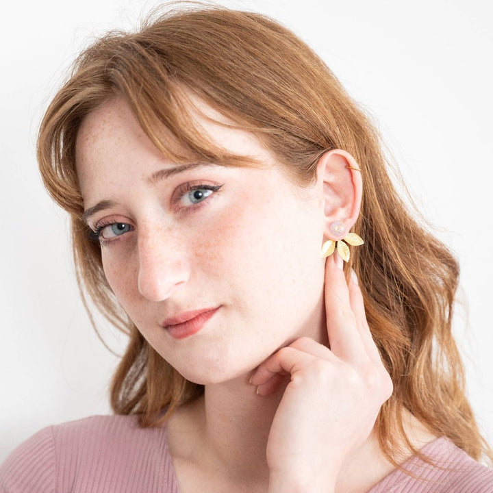 A pair of gold earrings with a leaf design. The earrings have a white, circular resin center with gold flecks. The leaves are attached to the center and can be detached.