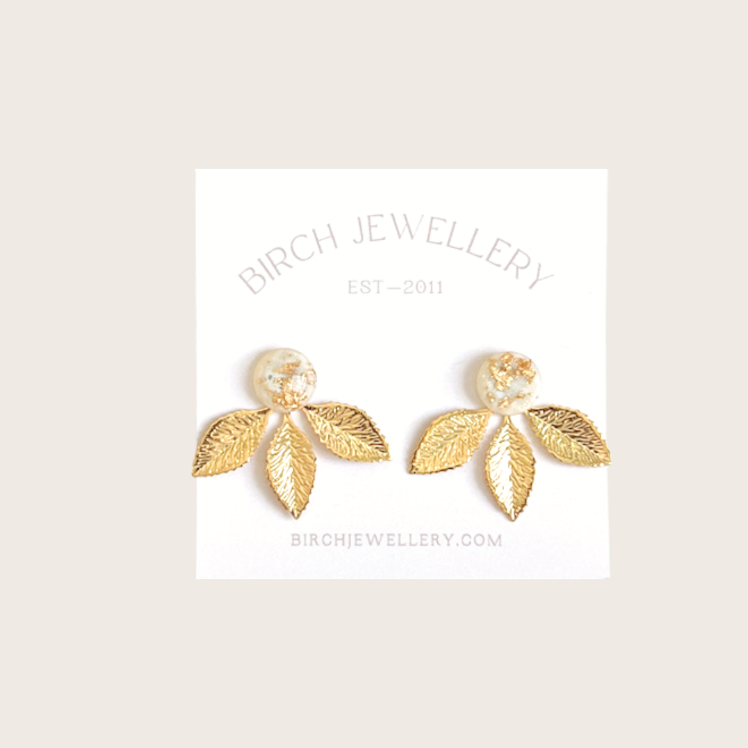 A pair of gold earrings with a leaf design. The earrings have a white, circular resin center with gold flecks. The leaves are attached to the center and can be detached.