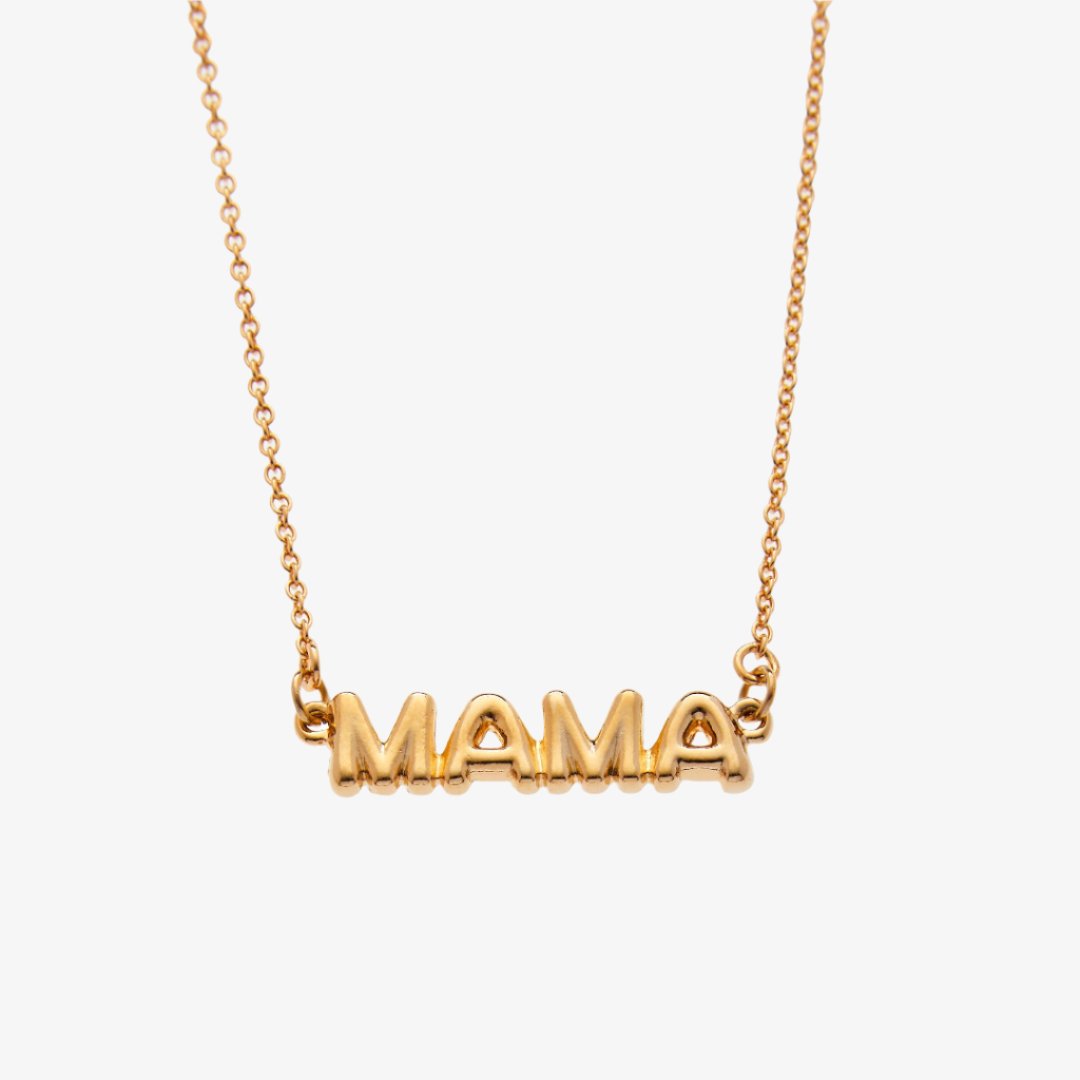 A gold necklace on a pink background. The necklace features a gold pendant with the word "MAMA" in bold letters. The text "COOL MAMA FOREVER" is printed on the background card, along with the brand name "Foxy Originals."