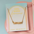 A gold necklace on a pink background. The necklace features a gold pendant with the word "MAMA" in bold letters. The text "COOL MAMA FOREVER" is printed on the background card, along with the brand name "Foxy Originals."