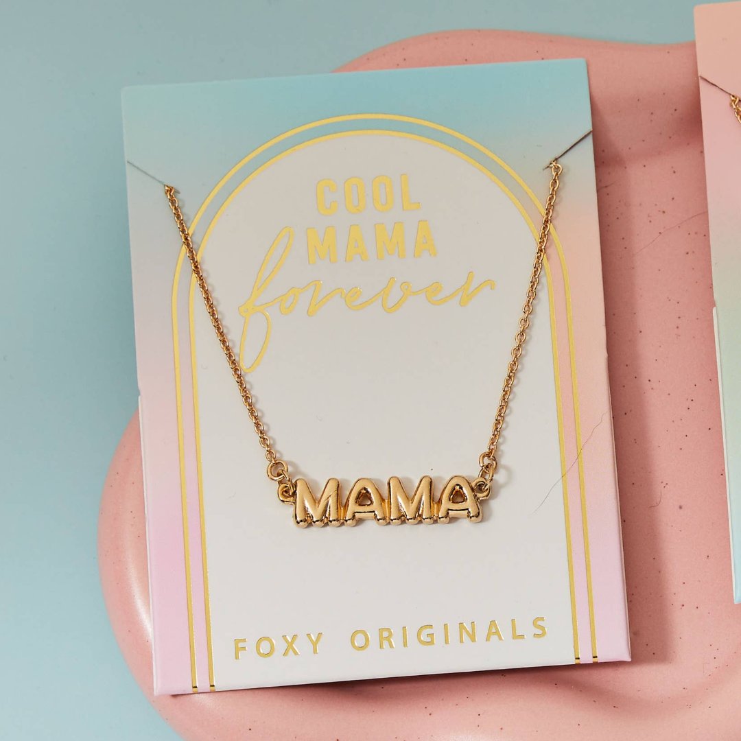 A gold necklace on a pink background. The necklace features a gold pendant with the word "MAMA" in bold letters. The text "COOL MAMA FOREVER" is printed on the background card, along with the brand name "Foxy Originals."
