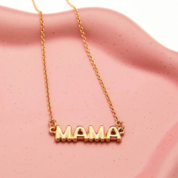 A gold necklace on a pink background. The necklace features a gold pendant with the word 