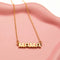 A gold necklace on a pink background. The necklace features a gold pendant with the word 