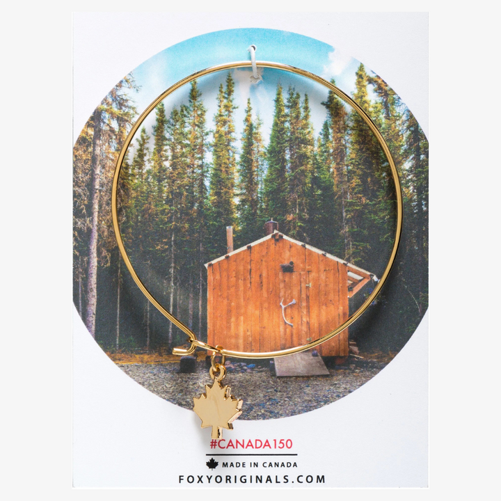 A gold bangle bracelet with a maple leaf charm. The bracelet is displayed on a card with a photo of a Canadian cabin in the woods and the hashtag #CANADA150. The card also includes the information 