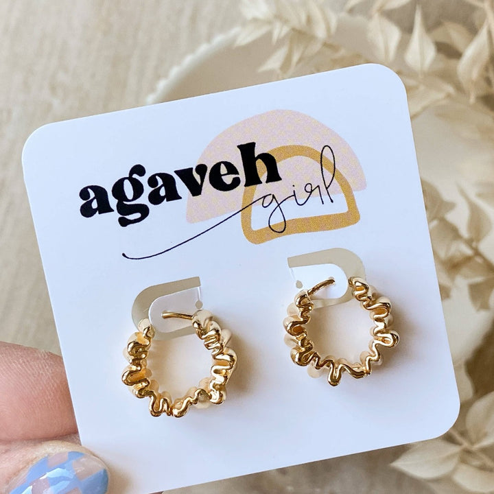 A pair of Agaveh Girl Gold Ribbon Hoops, featuring a 14k gold plated brass pleated lace design, displayed on a branded card with the text 'agaveh girl,' against a textured background with dried floral accents. The earrings are dainty and textured, available in Ottawa, Ontario, Canada.