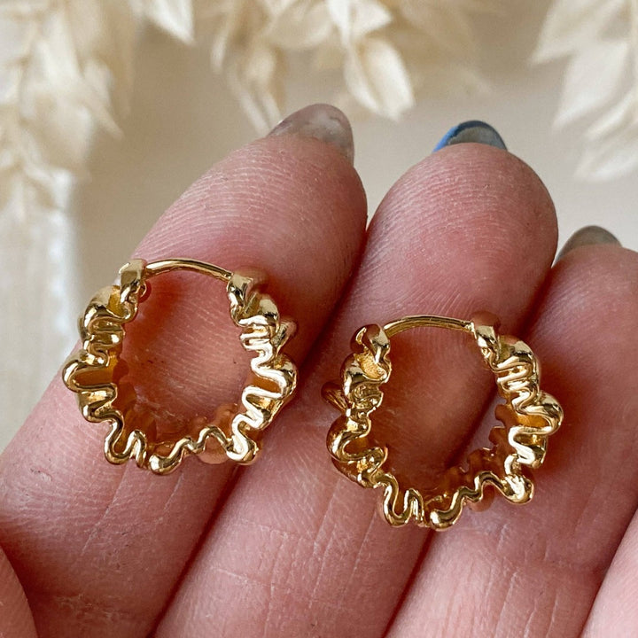 A pair of Agaveh Girl Gold Ribbon Hoops, featuring a 14k gold plated brass pleated lace design, displayed on a branded card with the text 'agaveh girl,' against a textured background with dried floral accents. The earrings are dainty and textured, available in Ottawa, Ontario, Canada.