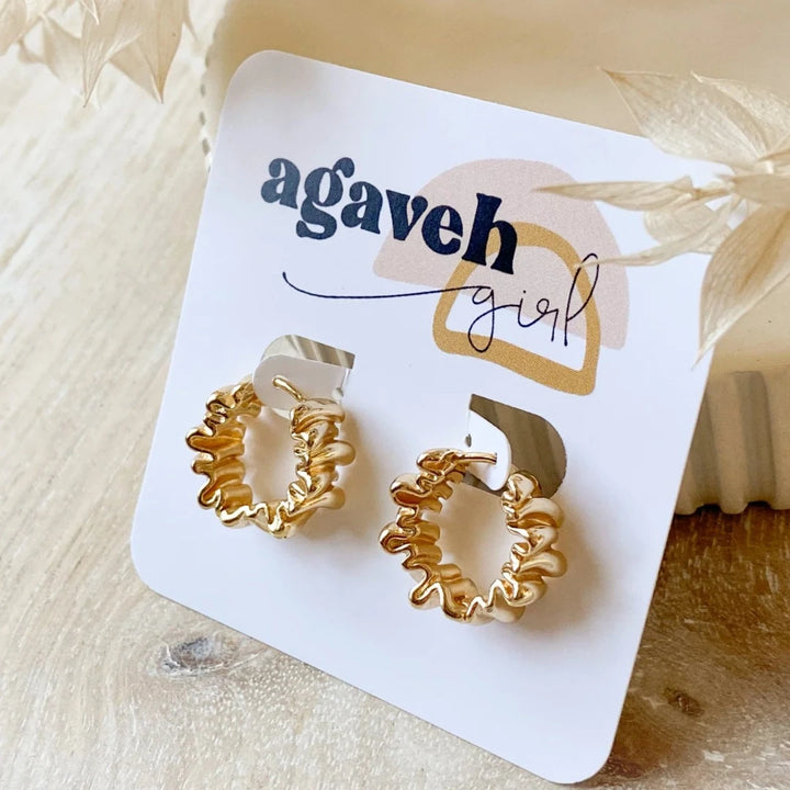 A pair of Agaveh Girl Gold Ribbon Hoops, featuring a 14k gold plated brass pleated lace design, displayed on a branded card with the text 'agaveh girl,' against a textured background with dried floral accents. The earrings are dainty and textured, available in Ottawa, Ontario, Canada.
