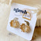 A pair of Agaveh Girl Gold Ribbon Hoops, featuring a 14k gold plated brass pleated lace design, displayed on a branded card with the text 'agaveh girl,' against a textured background with dried floral accents. The earrings are dainty and textured, available in Ottawa, Ontario, Canada.