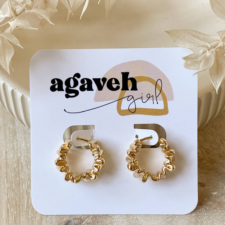 A pair of Agaveh Girl Gold Ribbon Hoops, featuring a 14k gold plated brass pleated lace design, displayed on a branded card with the text 'agaveh girl,' against a textured background with dried floral accents. The earrings are dainty and textured, available in Ottawa, Ontario, Canada.