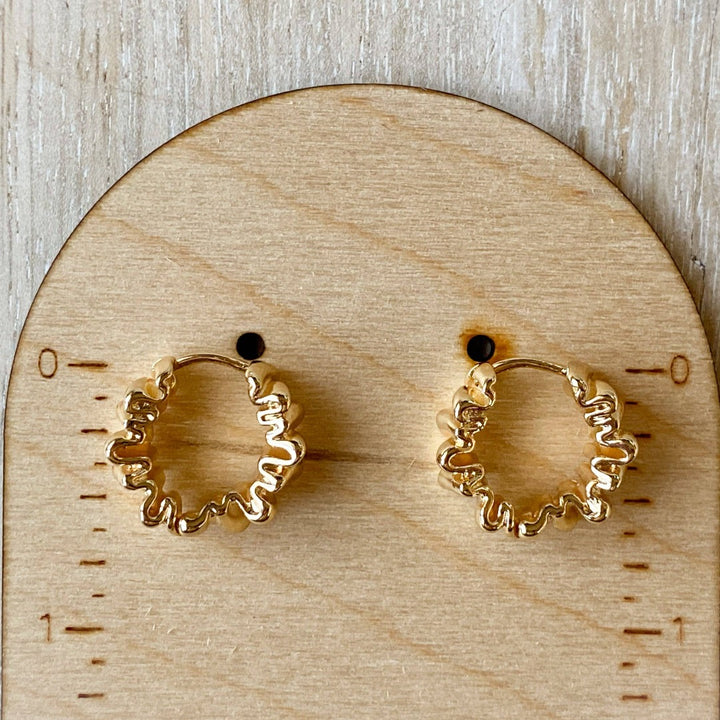 A pair of Agaveh Girl Gold Ribbon Hoops, featuring a 14k gold plated brass pleated lace design, displayed on a branded card with the text 'agaveh girl,' against a textured background with dried floral accents. The earrings are dainty and textured, available in Ottawa, Ontario, Canada.