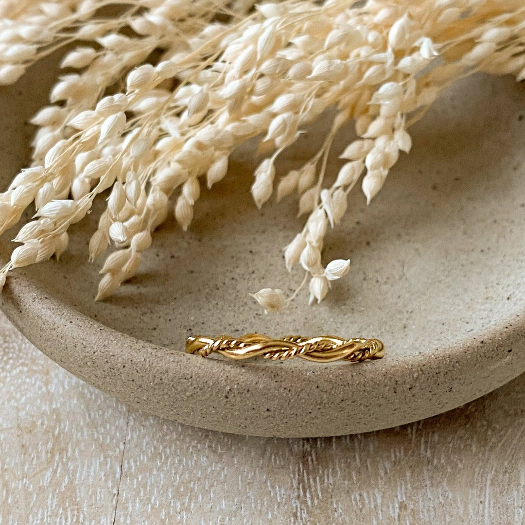 A gold twisted ring, featuring a woven or rope-like texture, displayed on a speckled ceramic dish with dried floral accents. The ring is dainty and stackable, available in Ottawa, Ontario, Canada.