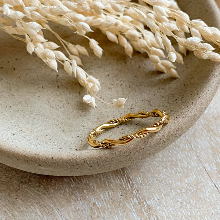 A gold twisted ring, featuring a woven or rope-like texture, displayed on a speckled ceramic dish with dried floral accents. The ring is dainty and stackable, available in Ottawa, Ontario, Canada.