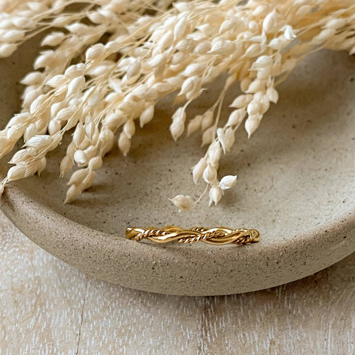 A gold twisted ring, featuring a woven or rope-like texture, displayed on a speckled ceramic dish with dried floral accents. The ring is dainty and stackable, available in Ottawa, Ontario, Canada.