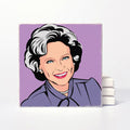 A square ceramic coaster featuring a stylized pop-art portrait of a smiling woman with short gray hair, blue eyes, and red lips, set against a lavender background. The coaster is displayed upright on a white surface with a stack of similar coasters behind it.