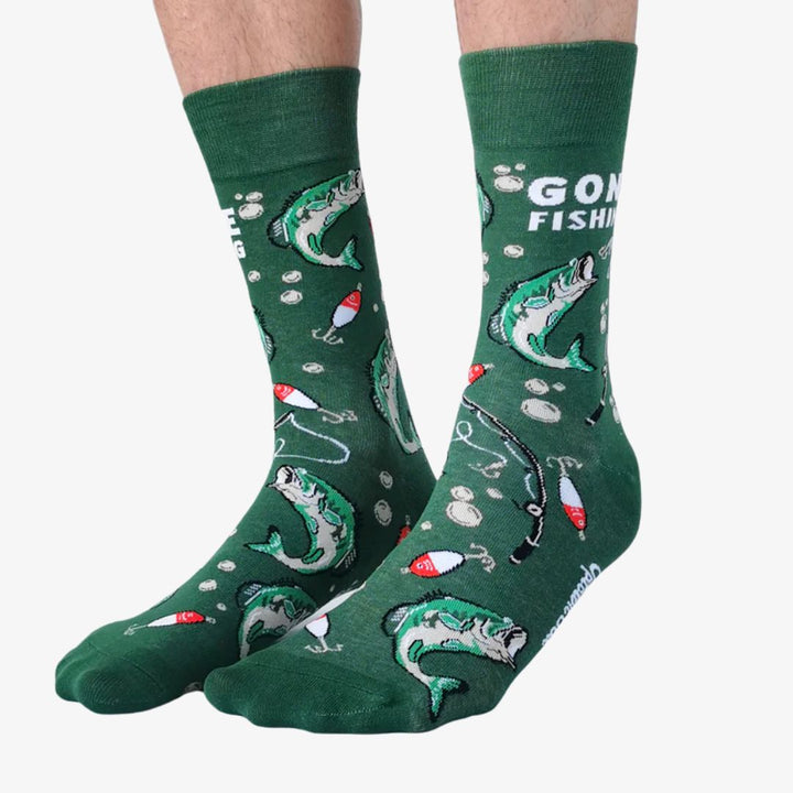 A pair of green crew socks featuring a fishing-themed pattern with illustrations of leaping fish, fishing rods, bobbers, and lures in shades of white, red, and black. One sock displays the text 