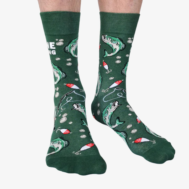 A pair of green crew socks featuring a fishing-themed pattern with illustrations of leaping fish, fishing rods, bobbers, and lures in shades of white, red, and black. One sock displays the text 