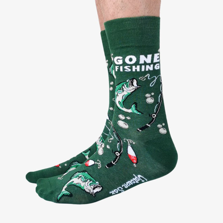 A pair of green crew socks featuring a fishing-themed pattern with illustrations of leaping fish, fishing rods, bobbers, and lures in shades of white, red, and black. One sock displays the text 
