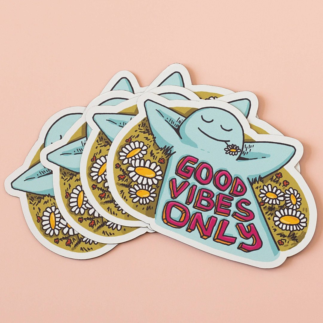 A blue, cartoon character with arms outstretched is lying on its back in a field of daisies. The text "GOOD VIBES ONLY" appears in bold, colorful letters below the character.